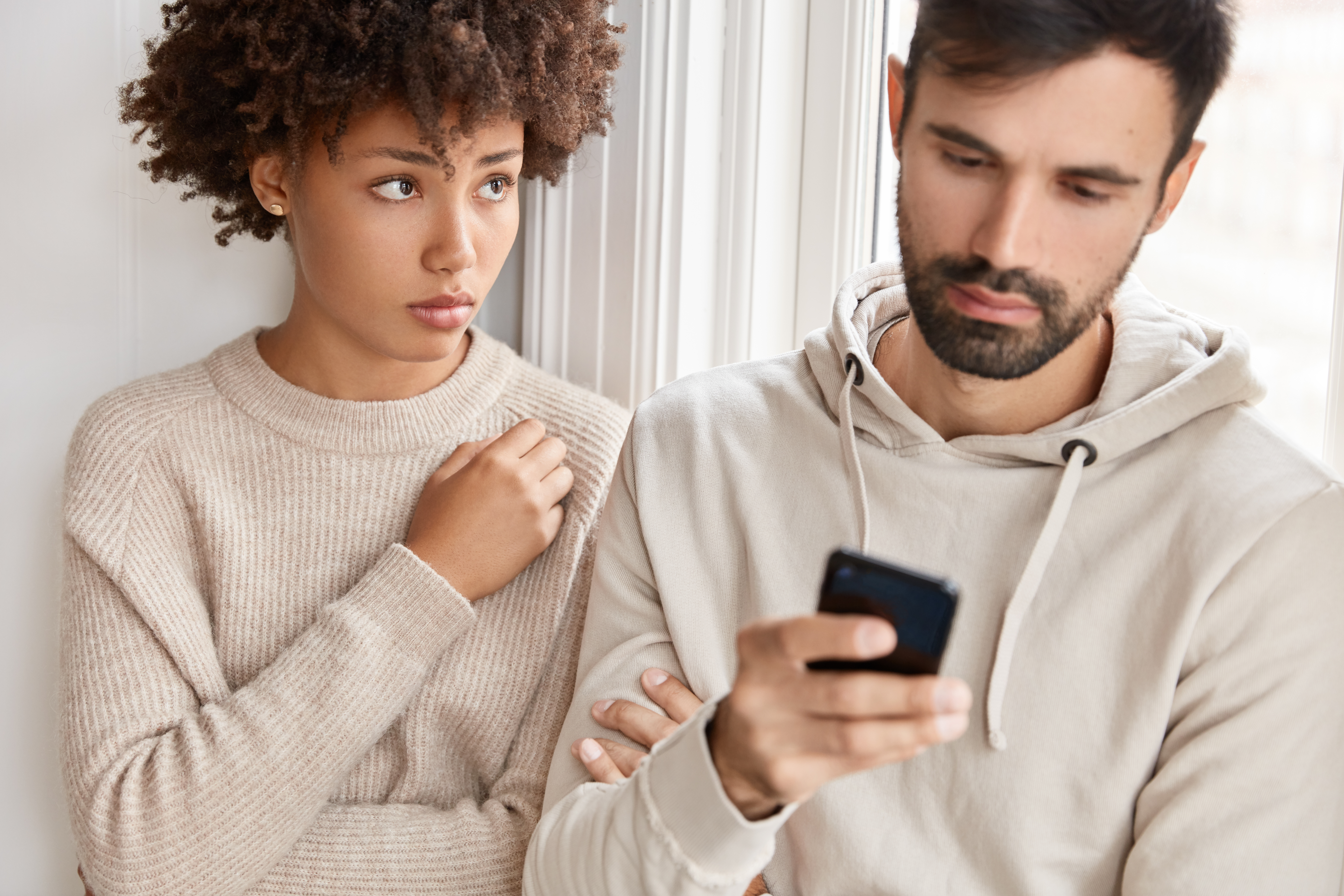 A woman appears upset while her partner looks at his phone privately, illustrating emotional infidelity and loss of trust.