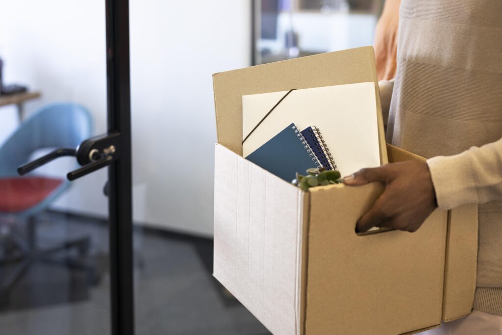 Close-up of hands holding a cardboard box with office items, calm office background—signaling a recent job transition.
